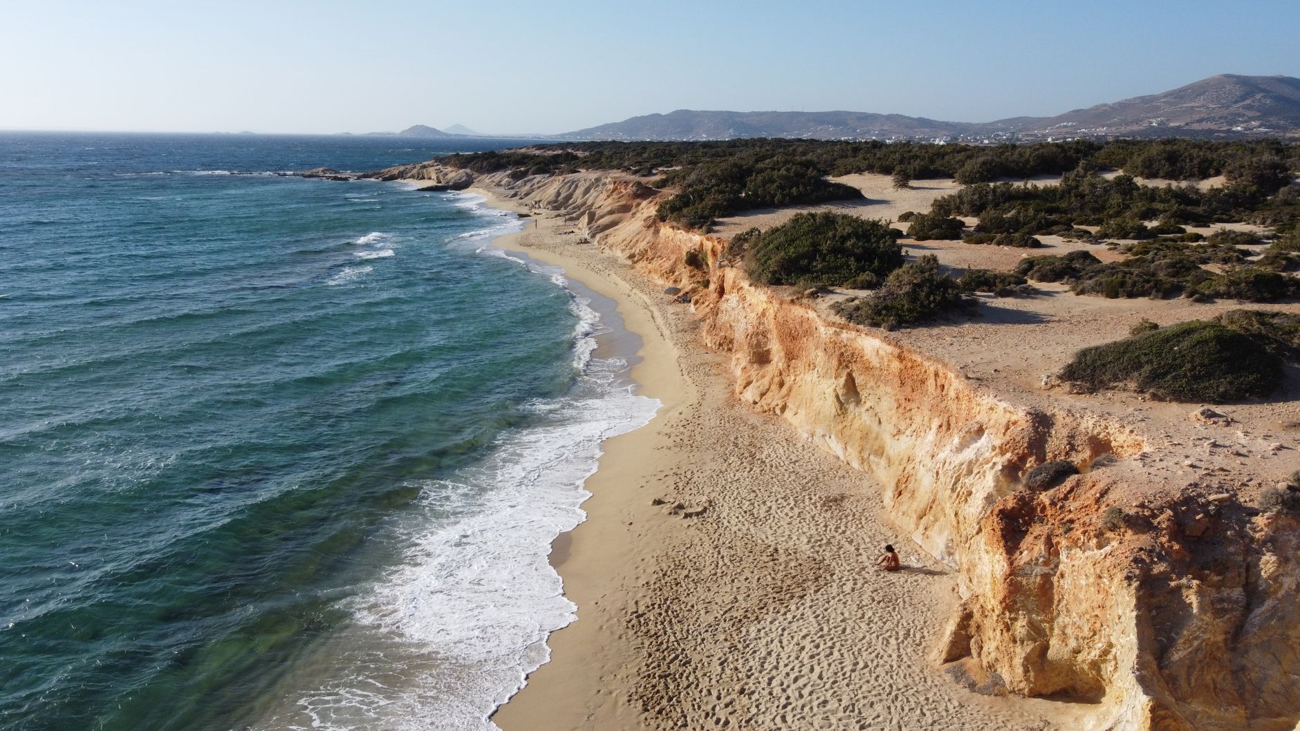Stunning aerial view of Naxos beach with rocky cliffs and clear blue sea under sunny skies in Greece.